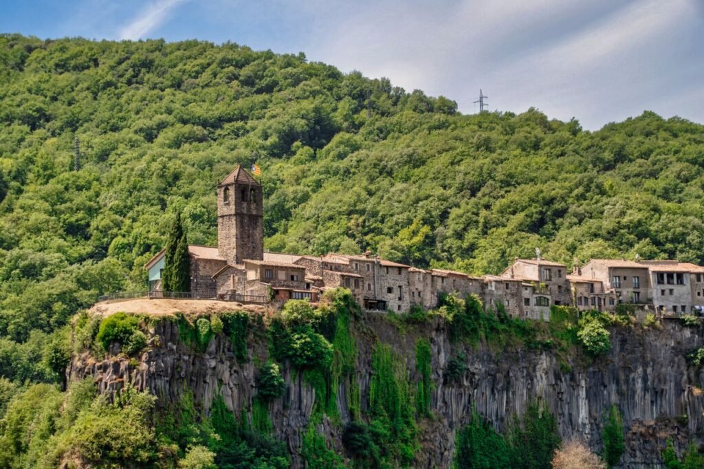 A scenic view of a medieval village perched on basalt cliffs with lush green forest in the background.