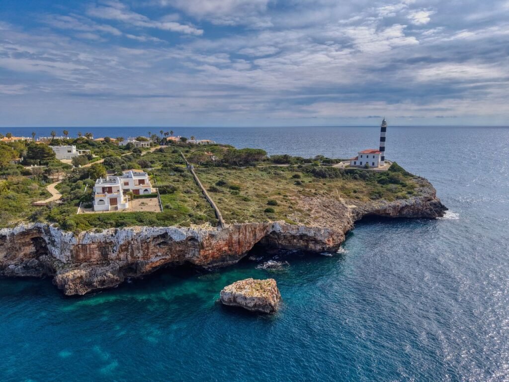 Scenic aerial view of a lighthouse on the rocky coast of Mallorca, Spain, under a partly cloudy sky.